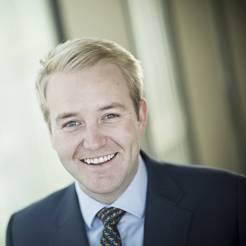 Smiling man in a suit and tie, standing indoors with soft lighting in the background.