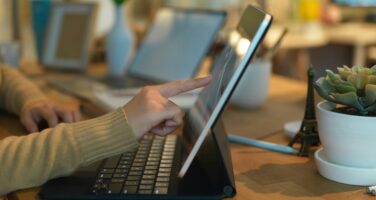 A person using a tablet with a keyboard and touching the screen on a wooden desk, surrounded by a plant and various office items.