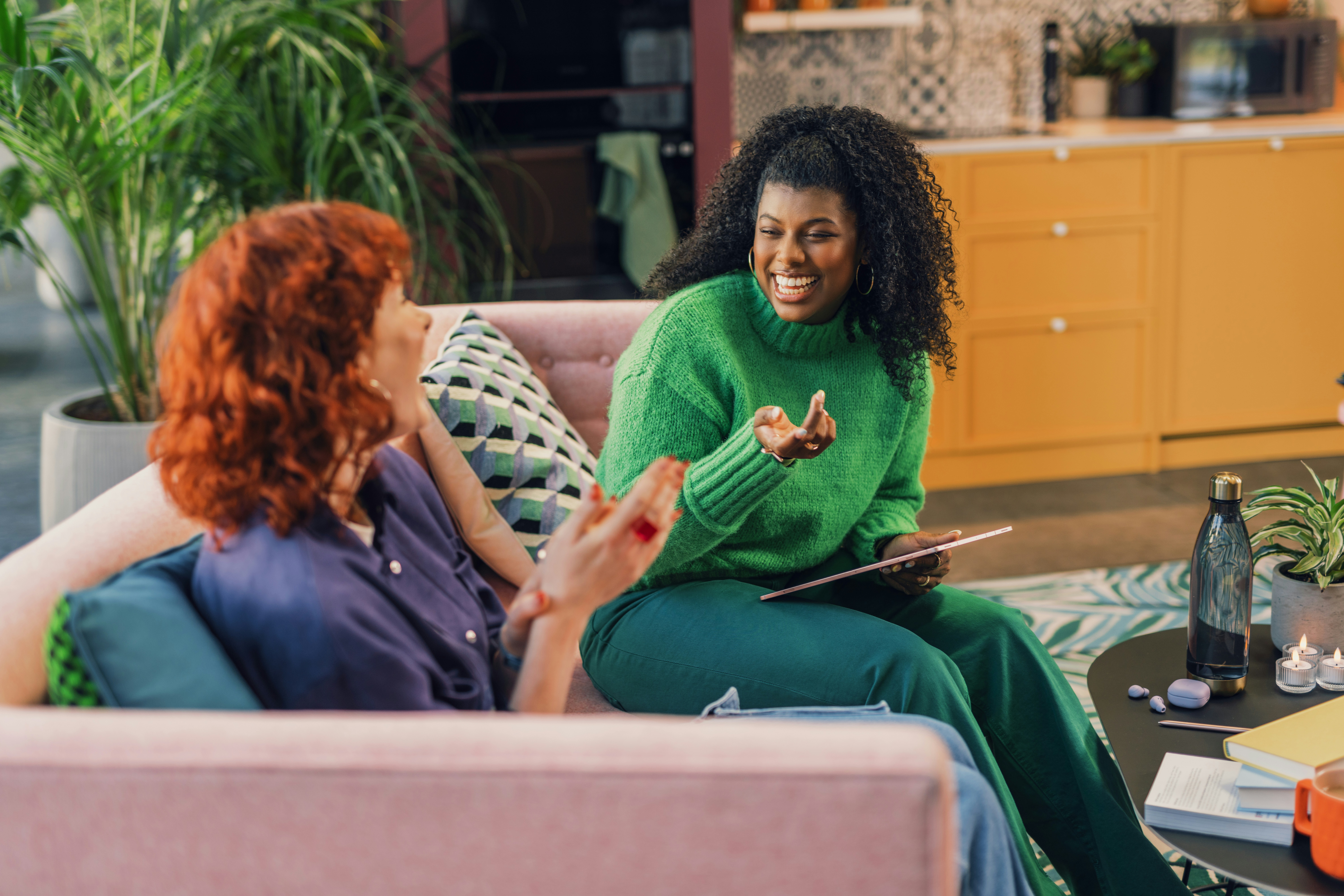 Two women are sitting on a sofa in a cozy living room, engaged in a friendly conversation. One woman is holding a tablet. The room is decorated with plants and has a warm, inviting atmosphere.