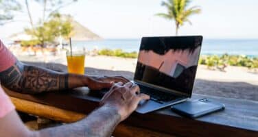 Person typing on a laptop at a beachside bar, with a tropical drink nearby and a view of the ocean and palm trees in the background.
