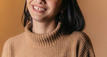 A smiling person with shoulder-length dark hair wearing a brown sweater against a warm, solid background.
