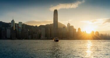 Sunset view of a city skyline with a tall building in the center, mountains in the background, and a boat on the water in the foreground.