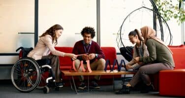 Four people sit around a table in a modern office. One person uses a wheelchair, and they engage with a tablet, appearing focused and collaborative.