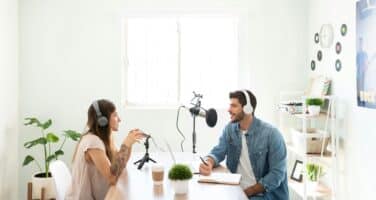 Two people wearing headphones are recording a podcast at a table with microphones. The room is bright and decorated with plants, shelves, and posters.