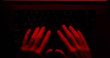Hands typing on a keyboard under red lighting, creating a dark and intense atmosphere.