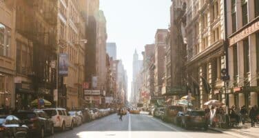 A sunlit urban street with parked cars on both sides, surrounded by tall buildings. A bicyclist rides down the center, and people walk along the sidewalks.