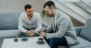 Two men in business attire sit on a sofa, engaged in a discussion. One is writing in a notebook, while the other listens attentively, both smiling. A table with two coffee cups is in front of them.
