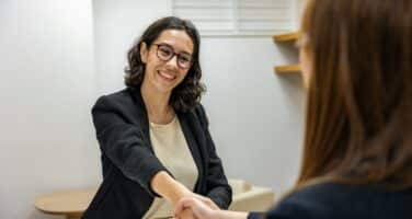 Two women in a professional setting shake hands across a table, one smiling and the other seen from behind.
