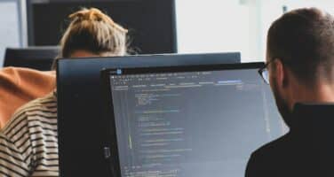 Two people working at computers in an office setting, focusing on code displayed on the screens.