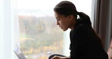 A woman sits in a chair by a window, focused on her laptop on a small table.