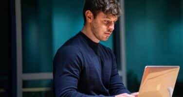 A man in a dark sweater is focused on working on a laptop at a desk in a dimly lit room.