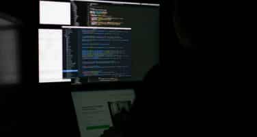 A person sitting in front of multiple computer screens displaying code in a low-light setting, suggesting a focus on programming or software development.