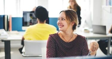 A woman in a patterned blouse smiles while sitting at a desk in a bright, modern office with people working in the background.