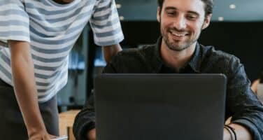 Two people smiling while looking at a laptop, suggesting a collaborative and positive work environment.