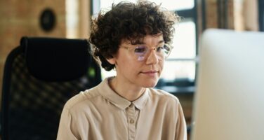 A person with curly hair and glasses is seated at a desk in an office, looking intently at a computer screen.