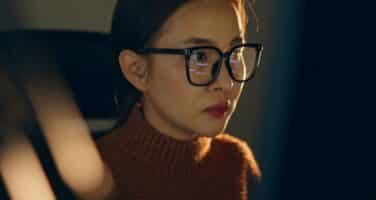 A woman wearing glasses focuses intently on a computer screen in dim lighting, reflecting determination and concentration.