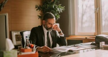 A man in a suit is sitting at an office desk, reading documents with a thoughtful expression.