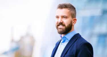 A bearded man in a blue suit smiles confidently while standing outdoors with a blurred modern glass building in the background.