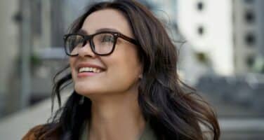 A woman with glasses and long dark hair smiles while looking up, standing outdoors in an urban setting.