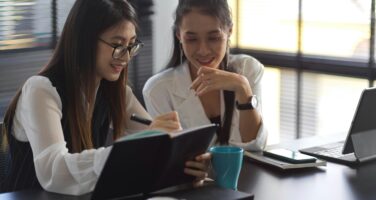 Two women in an office setting discuss work, one taking notes in a notebook. They are seated at a table with coffee cups, a laptop, and a smartphone.