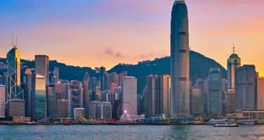 Hong Kong skyline at sunset with tall skyscrapers, including a prominent cylindrical building, set against a colorful sky and water in the foreground.