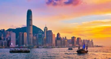 Hong Kong skyline at sunset with colorful sky, high-rise buildings, and boats on the water.
