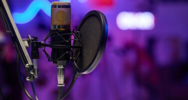 A studio microphone with a pop filter is mounted on a stand against a blurred purple and blue background, suggesting a professional audio or podcasting setup.