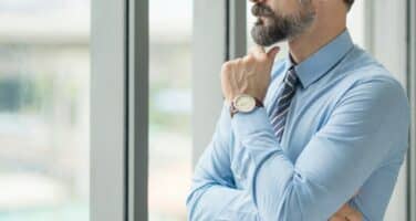 A bearded man in a blue shirt and tie stands near a large window, thoughtfully looking outside.