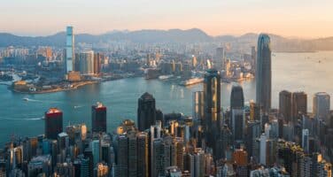 Aerial view of a cityscape with tall skyscrapers and a large body of water, surrounded by distant mountains under a clear sky during sunset.