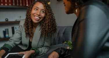 Two people are sitting and engaging in a friendly conversation. One person is holding a tablet and smiling. Shelves with books are visible in the background.