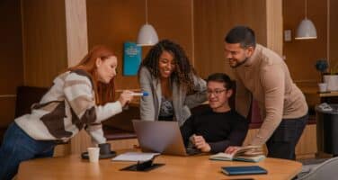 A diverse group of four people collaborates around a laptop in a modern office setting, discussing and smiling.