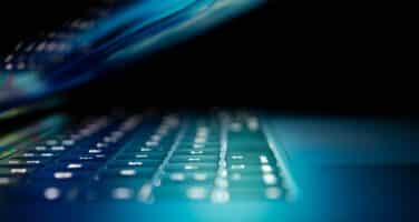 A partially open laptop with an illuminated keyboard emits a dim blue light in a dark environment.