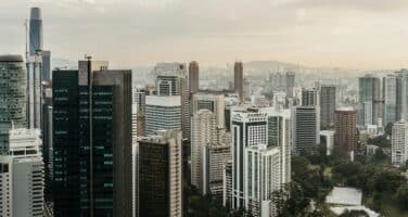 Aerial view of a modern city skyline with numerous high-rise buildings and a few green spaces, under a cloudy sky.