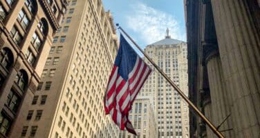 A United States flag hangs on a pole against a backdrop of tall, historic buildings under a blue sky in a cityscape.