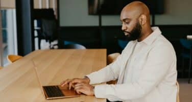 A man in a white shirt types on a laptop at a wooden table in a modern office setting, with a smartphone nearby.