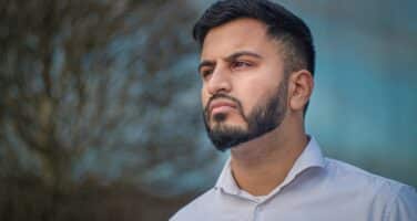 A man with a beard and short hair is looking thoughtfully into the distance, standing outdoors with a blurred tree and blue sky in the background.