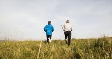 Two people jogging uphill on a grassy field under a clear sky, wearing blue and white jackets, respectively.