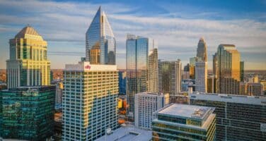 Skyline of a modern city with tall, reflective skyscrapers under a clear blue sky, forming a bustling urban landscape at sunset.