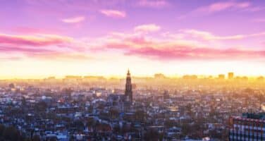 A cityscape at sunset with a silhouette of a central tower, vibrant purple and pink clouds, and buildings below.
