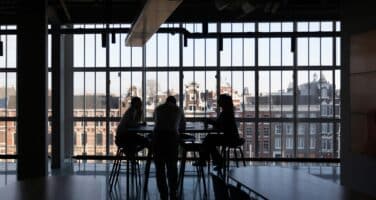 Three people sit around a table in silhouette against large windows, with an urban cityscape visible outside.