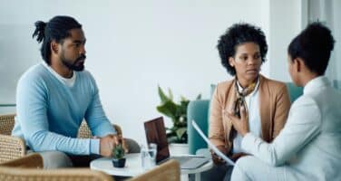Three people are engaged in a serious discussion in a modern office setting, with a laptop on the table and plants in the background.
