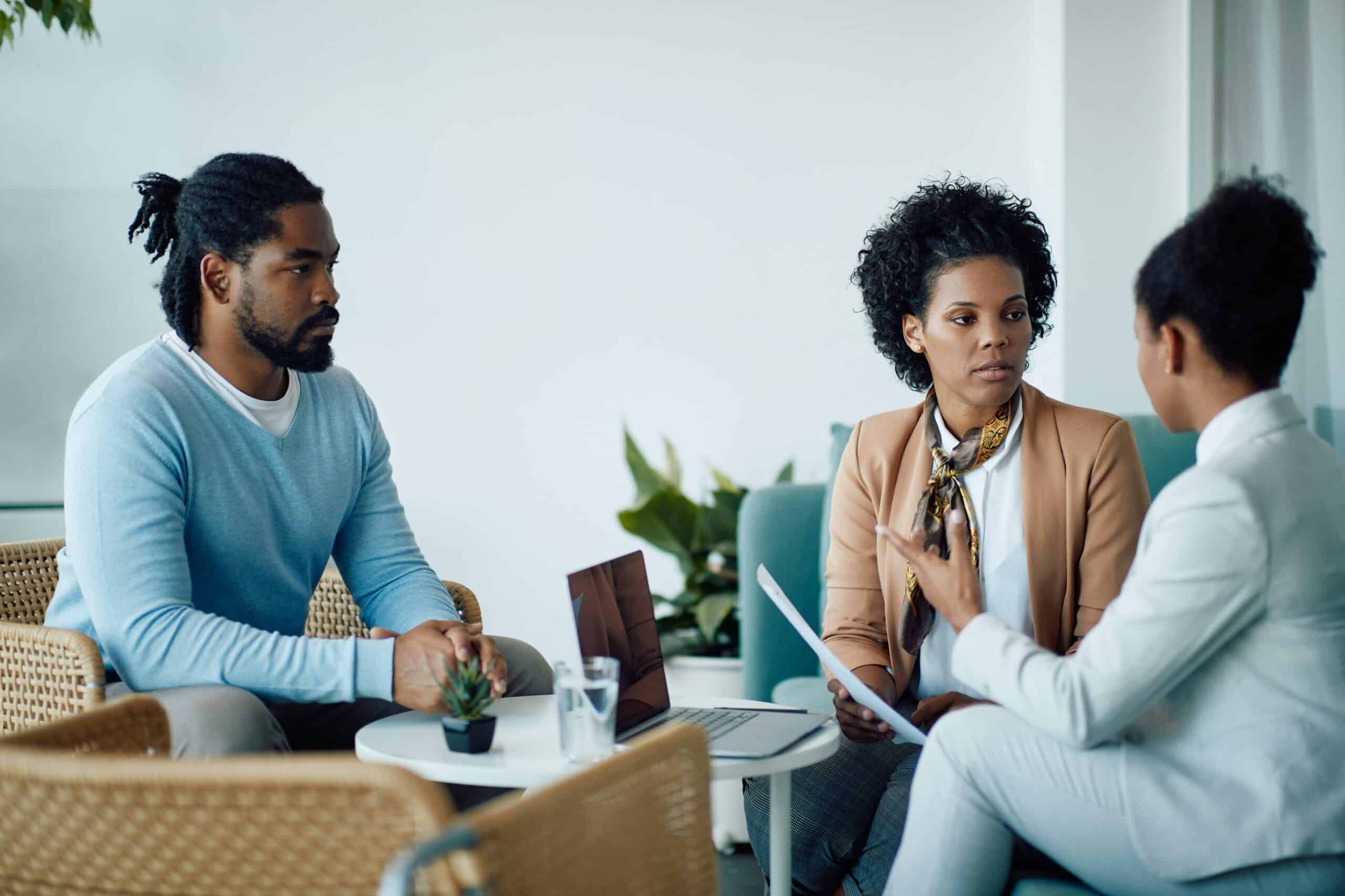 Three people are engaged in a serious discussion in a modern office setting, with a laptop on the table and plants in the background.