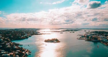 Aerial view of a calm coastal landscape with scattered islands, illuminated by sunlight. The sky is partly cloudy, reflecting on the tranquil blue water, creating a serene atmosphere.