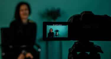 A woman is sitting in front of a camera being filmed, with her image visible on the camera's screen.