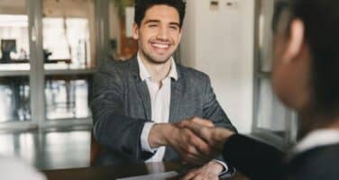 A smiling man in a suit shakes hands with someone across a table, suggesting a successful job interview or business meeting in a modern office setting.
