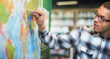 A person in glasses and a checkered shirt points at Europe on a world map in a library setting.