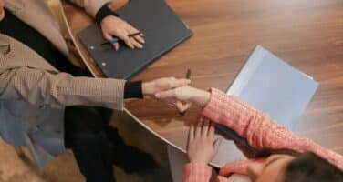Two people shaking hands across a wooden table with documents and notebooks nearby, suggesting a business meeting or agreement.