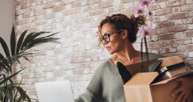 A woman in glasses holds a cardboard box with a potted orchid while looking at a laptop.