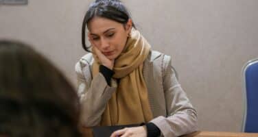 A woman wearing a scarf sits at a wooden table, looking thoughtful. She has a closed laptop in front of her. Another person is partially visible in the foreground.