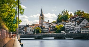 View of a tranquil river with a bridge leading to a picturesque European town, featuring classic buildings and a prominent clock tower under a clear sky.